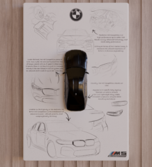 Top-view 3D-printed BMW M5 CS miniature in gloss-black finish mounted on a white panel with sketch background